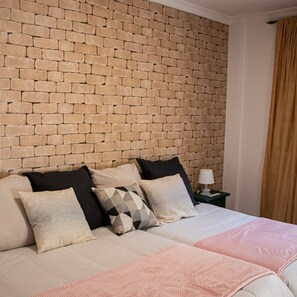 Una habitación de hotel con cabecera de ladrillo, cama con varias almohadas, mesita de noche con lámpara y ventana con cortinas.