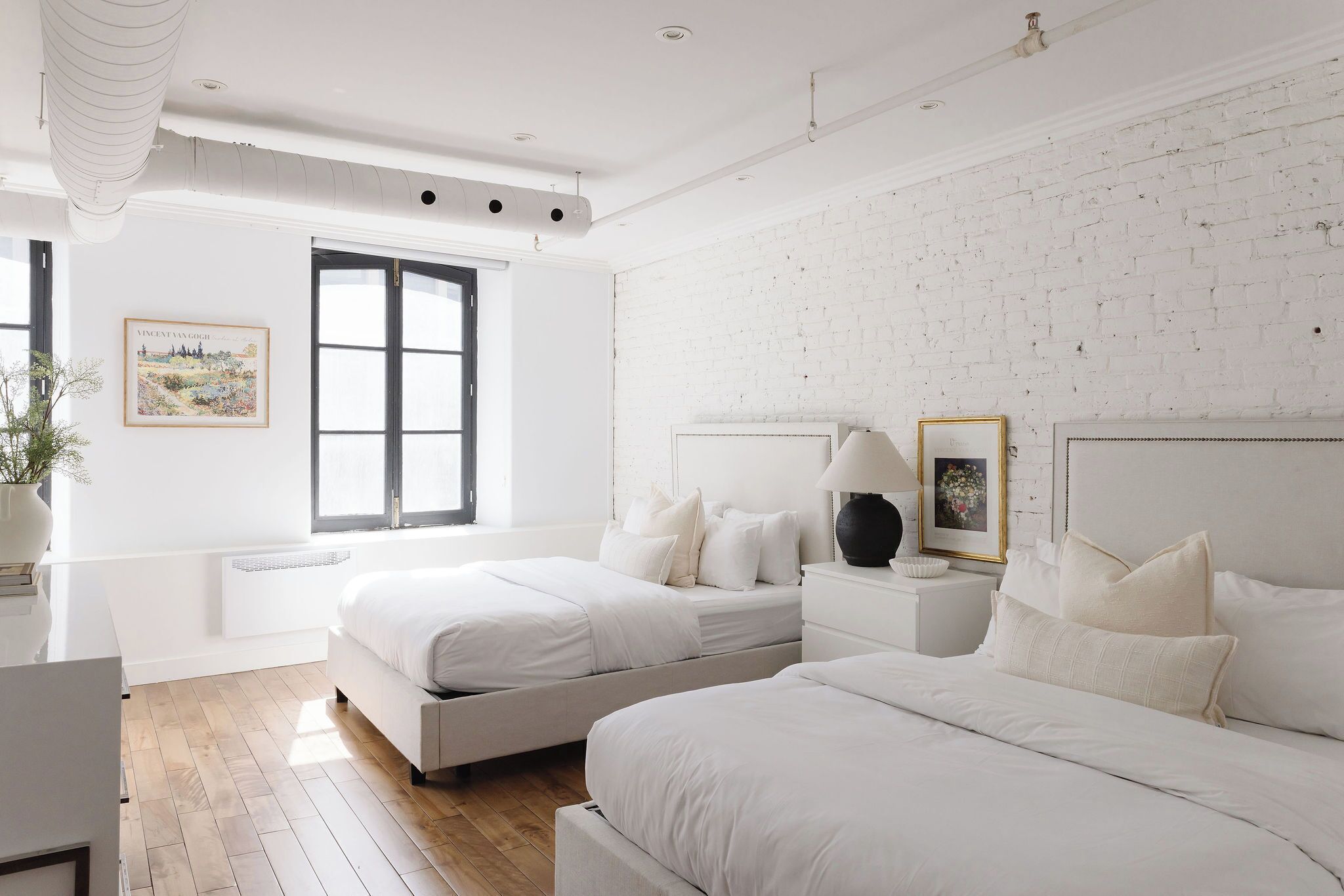 A modern bedroom with two beds, a white brick wall, and a large window.