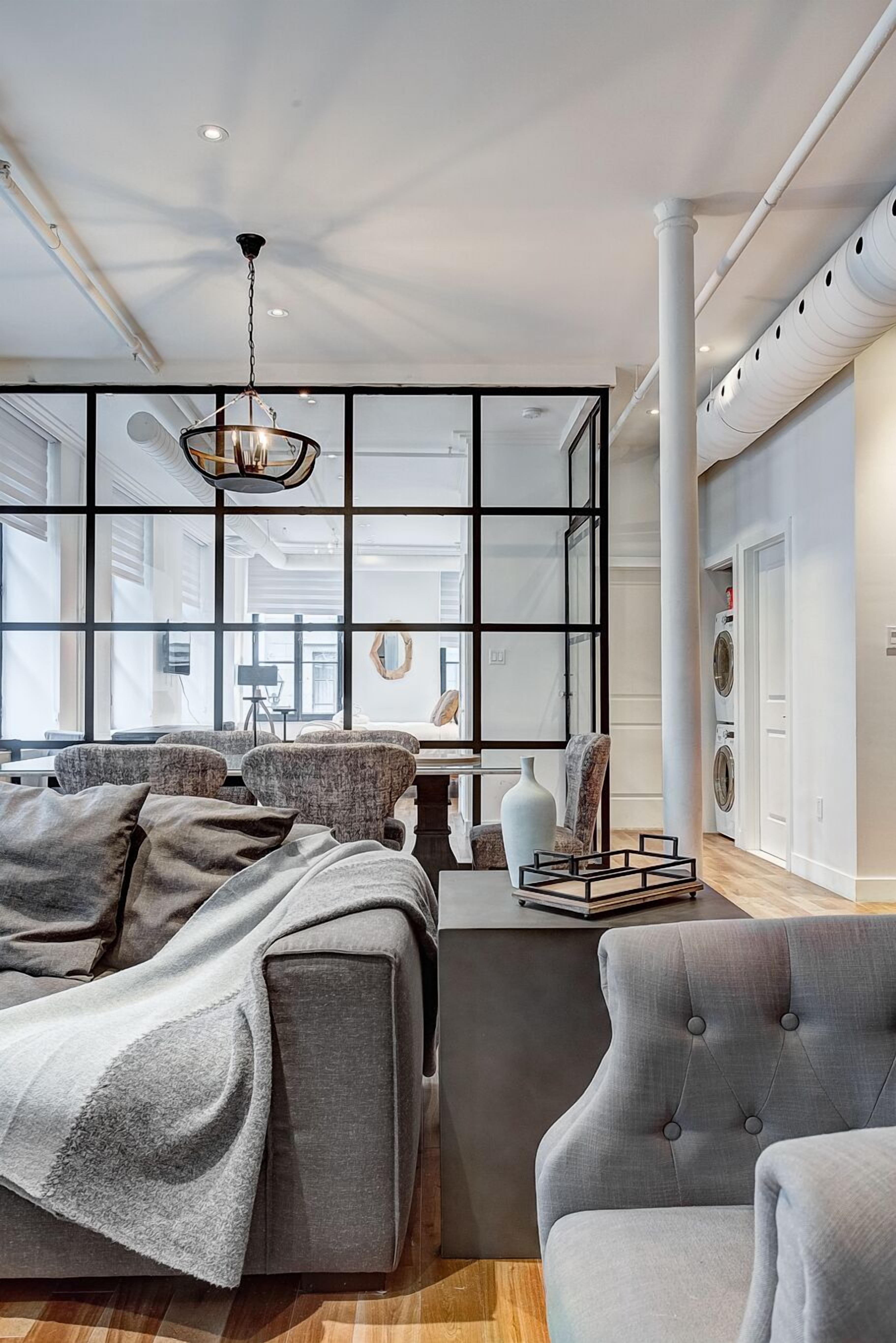 A modern bedroom with a large bed, a seating area with a chair, a hanging light fixture, and a view through a glass partition.