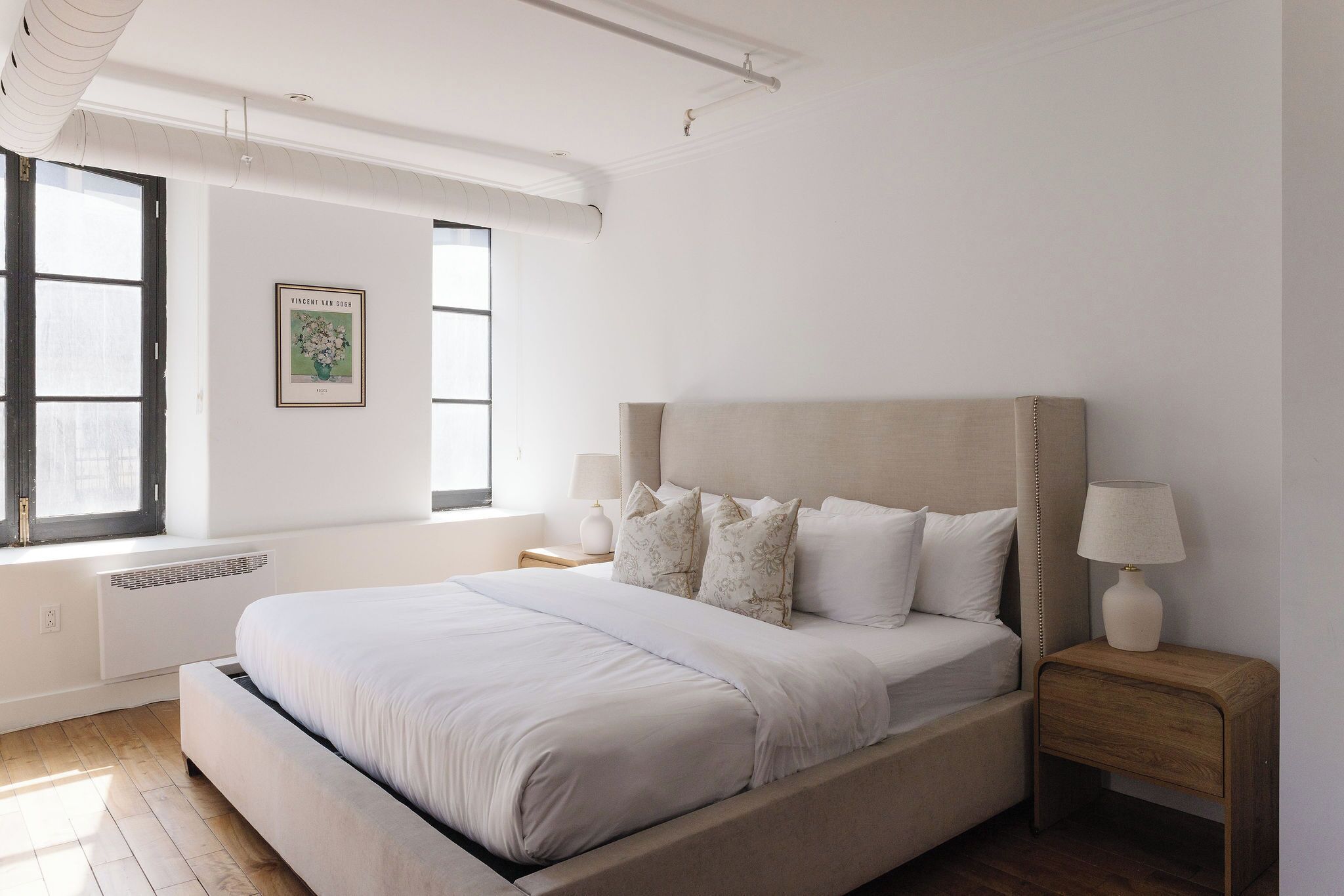 A bedroom with a bed, bedside tables, a radiator, a window, and a wall-mounted picture.
