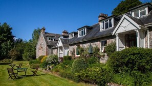 A stone building with a cross on the front, a lawn with chairs, and a garden with various plants. - The Cnoc Hotel (Beauly)