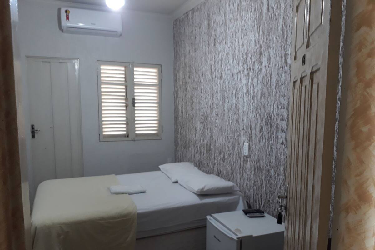 Habitación pequeña con cama individual, mesita de noche, aire acondicionado de pared y ventana con contraventanas.
