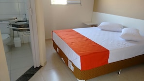 A hotel room with a bed, a small bedside table, a window, and a bathroom visible through an open door. - Hotel Casas Novas (Penapolis)