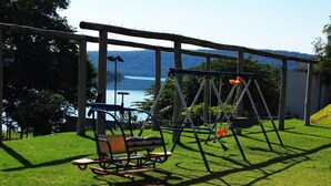 A swing set and a rocking chair in a grassy area with a view of a lake and mountains. - Enseada Nautico - JC Temporada (Caldas Novas)