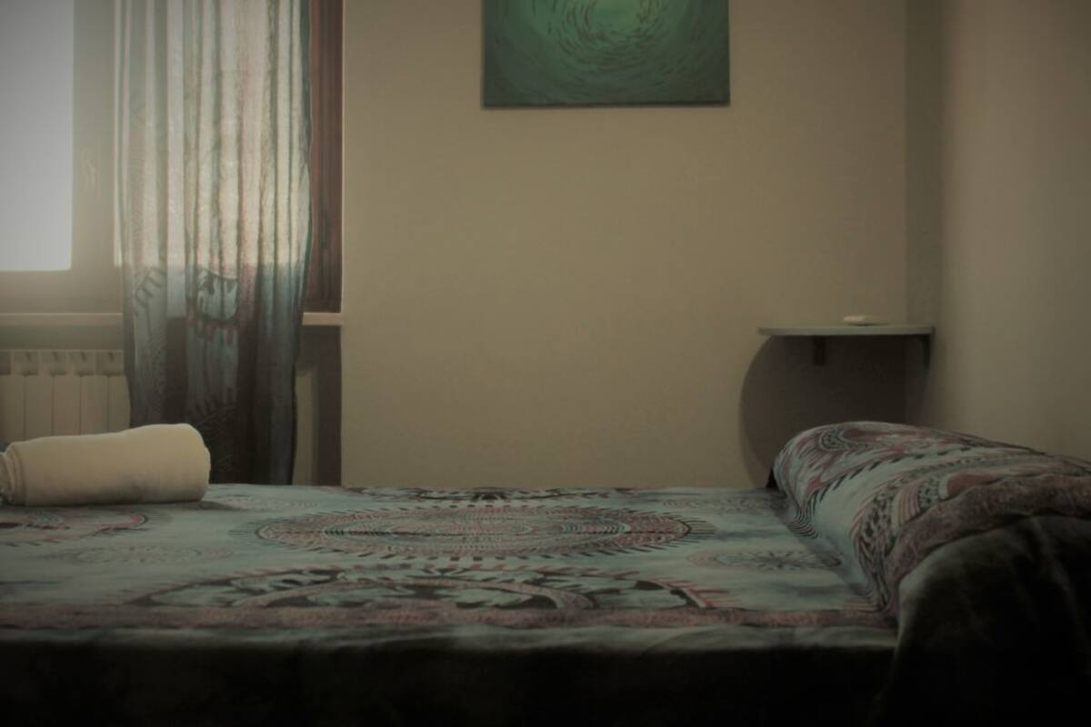 A bedroom with a bed, a window with curtains, a wall-mounted artwork, and a bedside table.