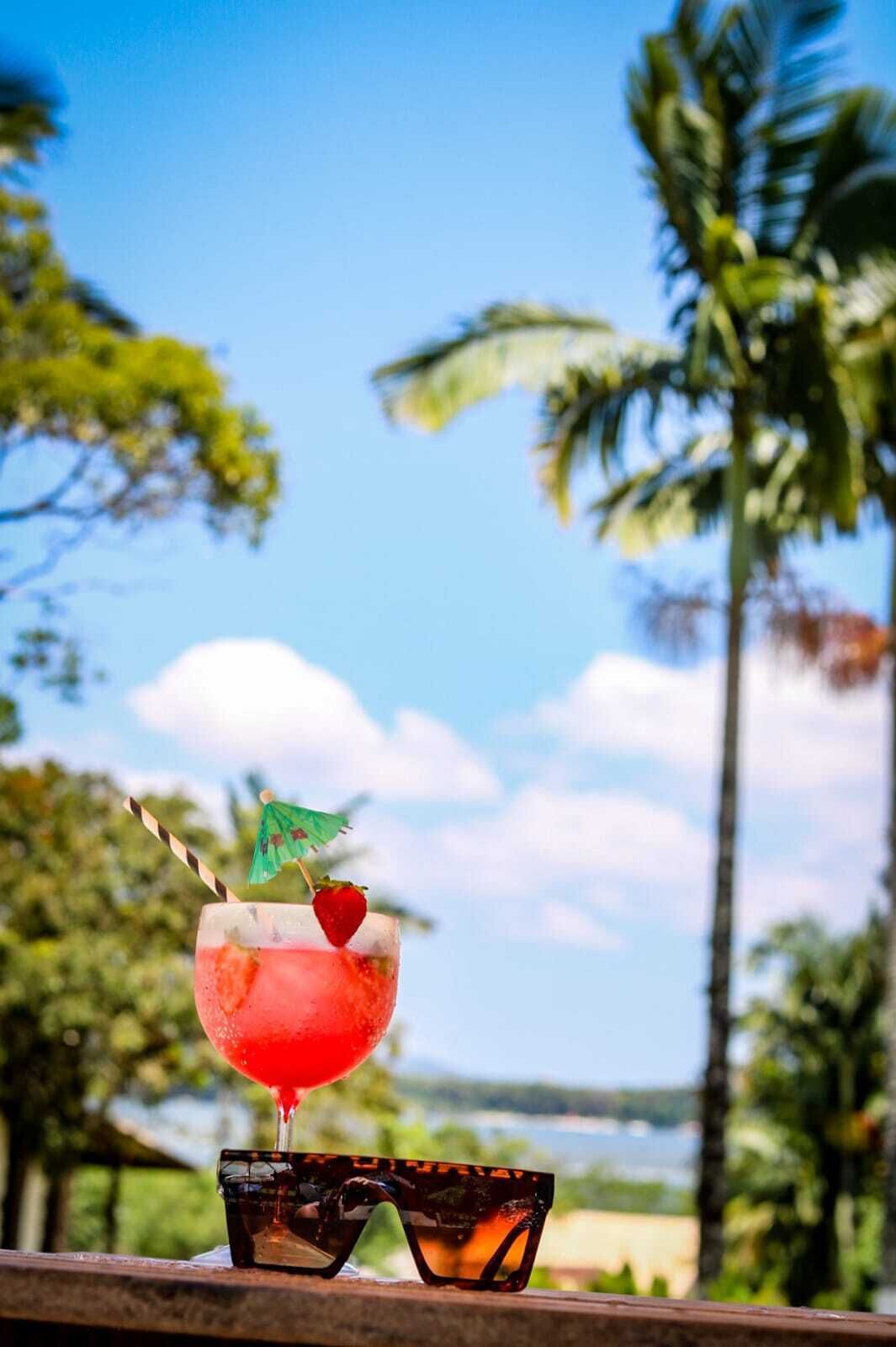 A tropical drink with a straw and a cherry on a glass, placed on a wooden surface with sunglasses beside it, set against a backdrop of palm trees and a clear sky.