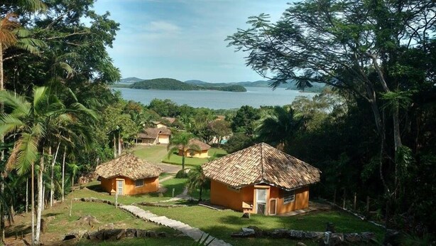 A tropical resort with thatched-roof huts surrounded by lush greenery and a view of a calm lake. - Pousada Villa da Glória (São Francisco do Sul)