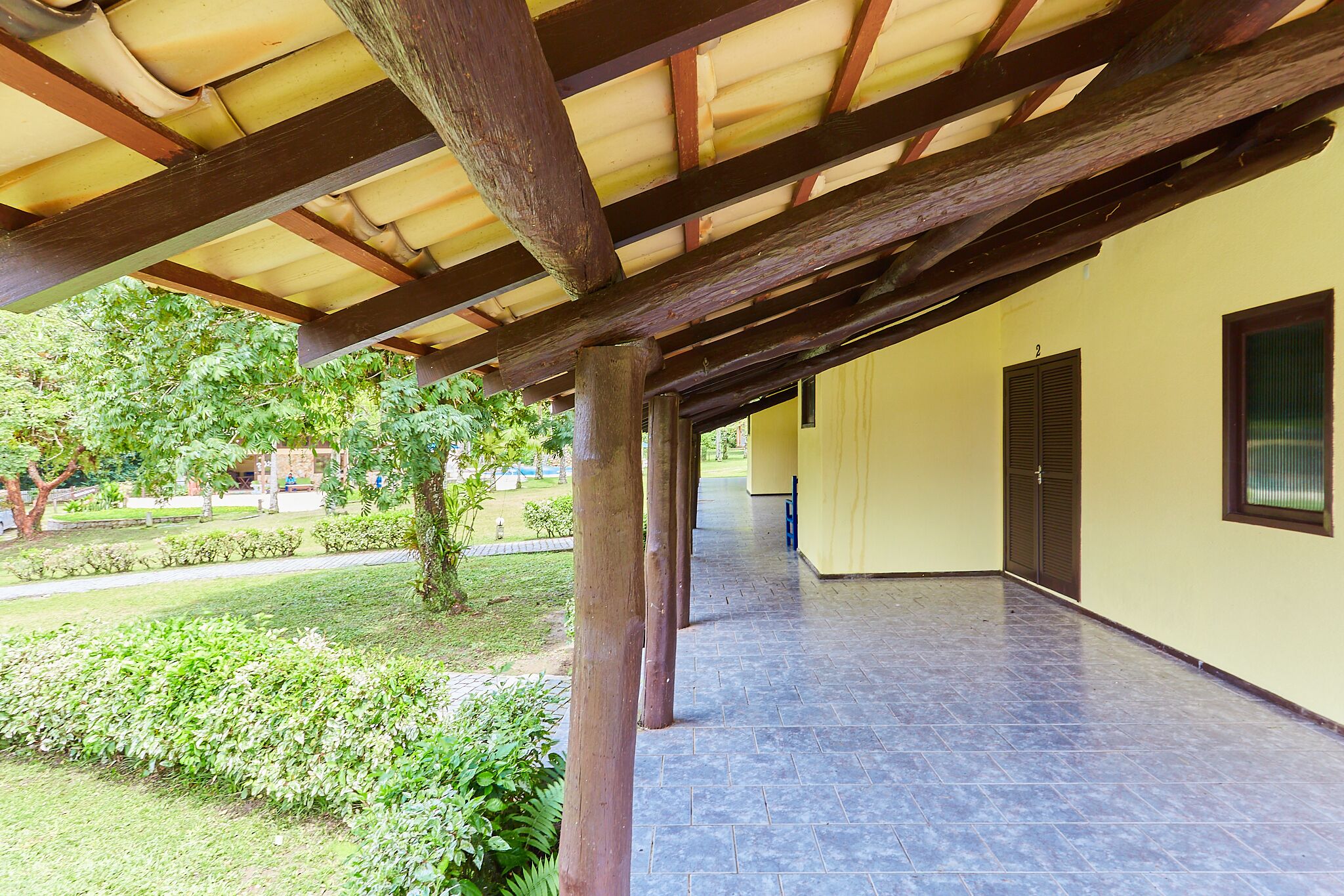 A covered outdoor area with a tiled floor, wooden beams, and a yellow wall with a window and door.