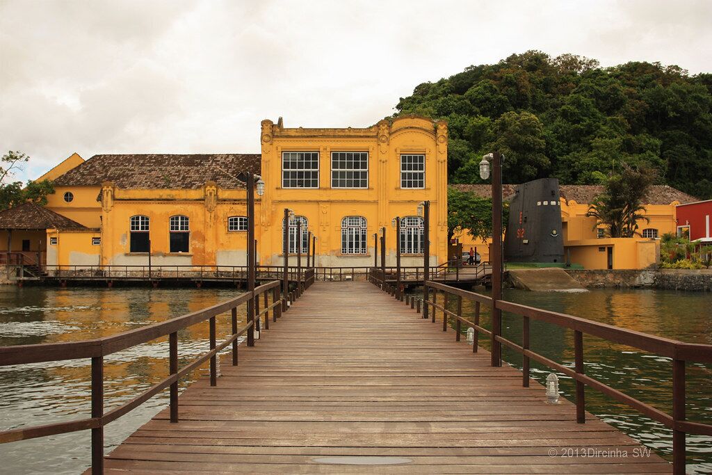 A wooden pier leading to a yellow building with multiple windows and a balcony, surrounded by water and greenery.