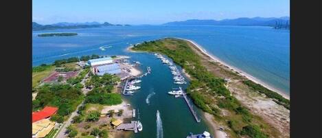 An aerial view of a coastal marina with boats docked along the shore, a pier extending into the water, and a building complex with a red roof.