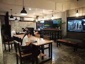 A restaurant with wooden tables and chairs, pendant lighting, and a large window with a view of the street. - OasisCafe&Nap - Hostel (Bangkok)