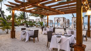 An outdoor dining area with tables set for two, under a wooden pergola with string lights, overlooking a beach with palm trees and a view of the sea. - Be Live Collection Punta Cana - Adults Only (Punta Cana)