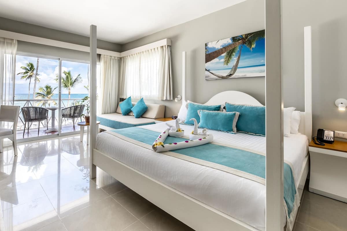 A modern hotel room with a large bed, a desk, a chair, and a balcony with a view of the ocean.