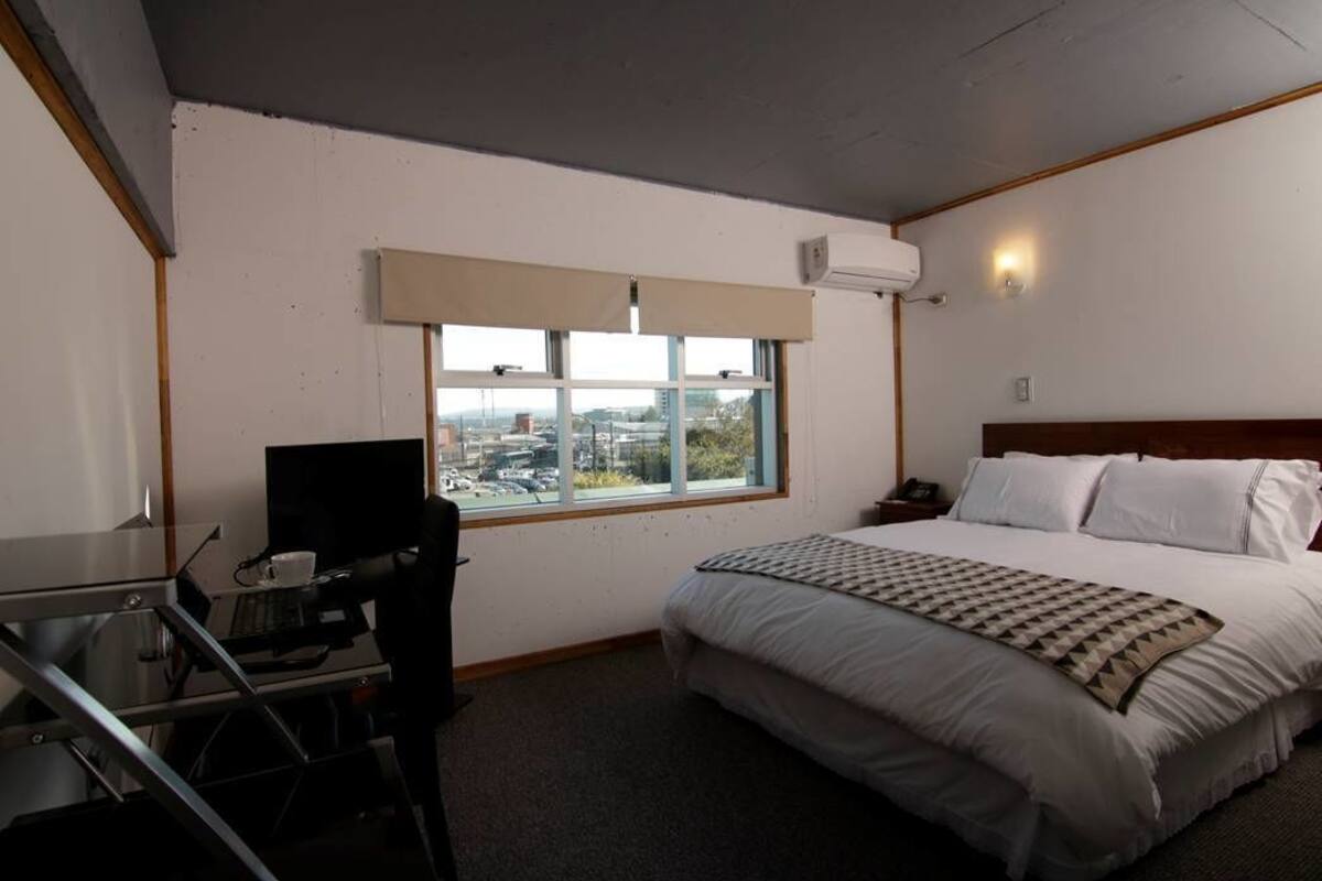 A hotel room with a bed, a desk with a computer, a chair, a window with a view of the city, and an air conditioning unit.