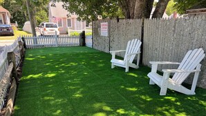 A fenced backyard with artificial grass, two Adirondack chairs, and a wooden fence. - Doryman Motel (Dennis Port)