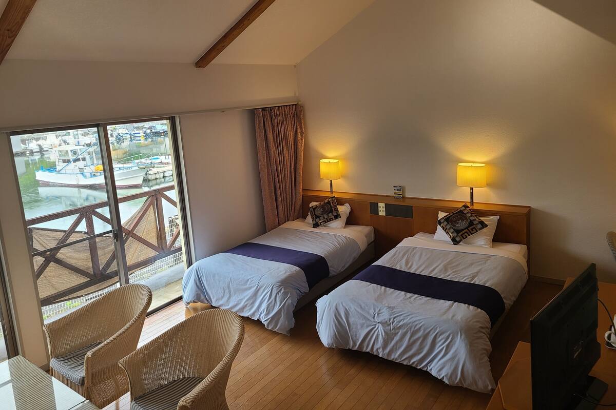 A hotel room with two beds, a balcony, wicker chairs, and a television.