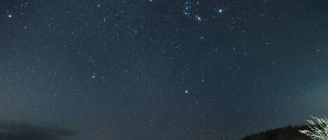 A night sky filled with stars over a coastal area with a distant mountain and a lit structure on the right.