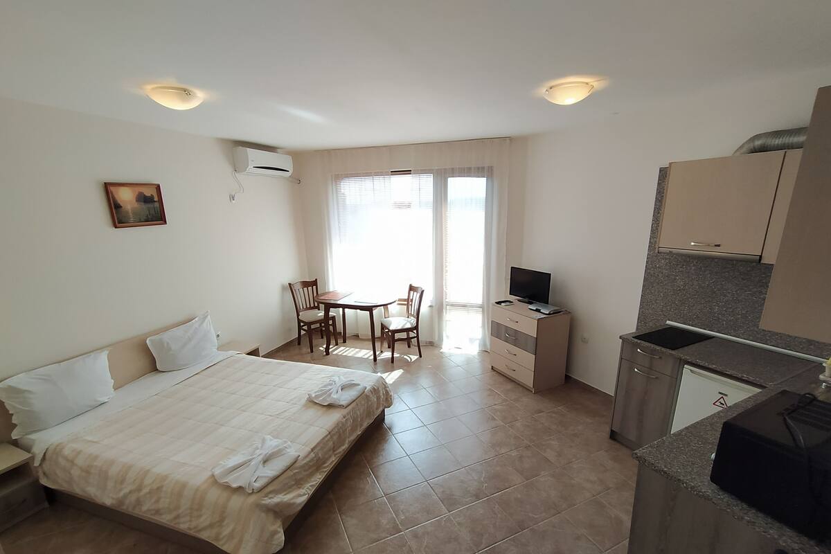 A hotel room with a bed, a desk, a chair, a television, a kitchen area, and a window with curtains.