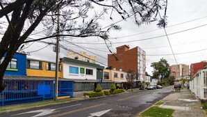 A street view with a blue building, a yellow building, and a red building. There are cars parked on the street and a sidewalk with a tree in the foreground.