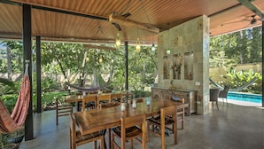 An open-plan dining area with a wooden table and chairs, a hammock, and a view of a swimming pool. - Villas Piña - Adults Only (Cahuita)