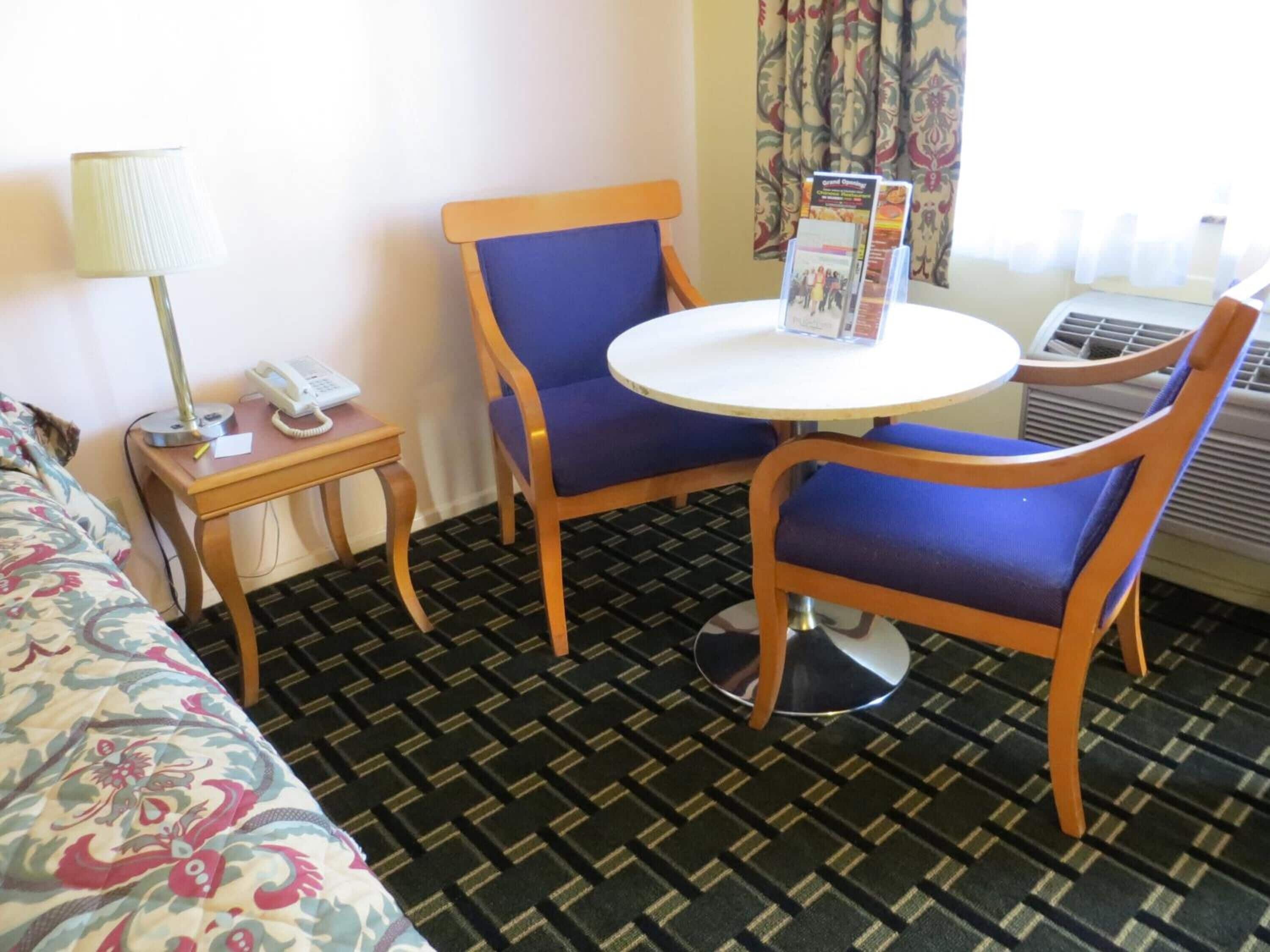 Habitació d'hotel amb un llit, una taula petita amb una làmpada, dues cadires, una taula rodona amb fulletons i un aparell d'aire condicionat.