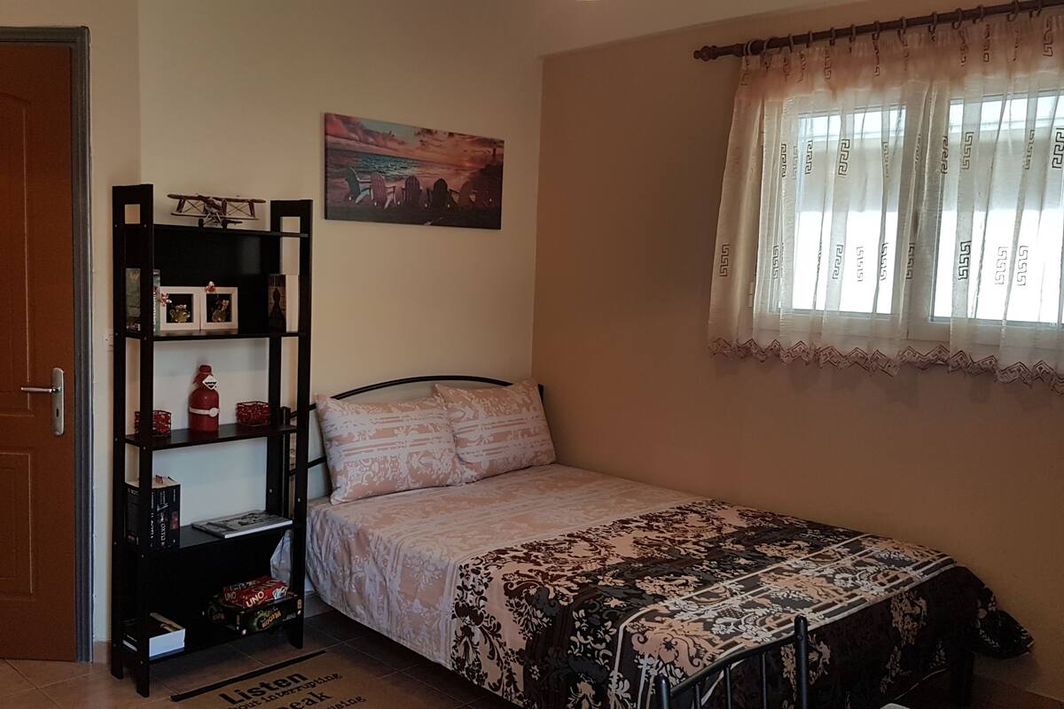 Ein Schlafzimmer mit einem Bett, einem Regal mit Büchern und einem kleinen Flugzeugmodell, einem Bild an der Wand und einem Fenster mit Vorhängen.