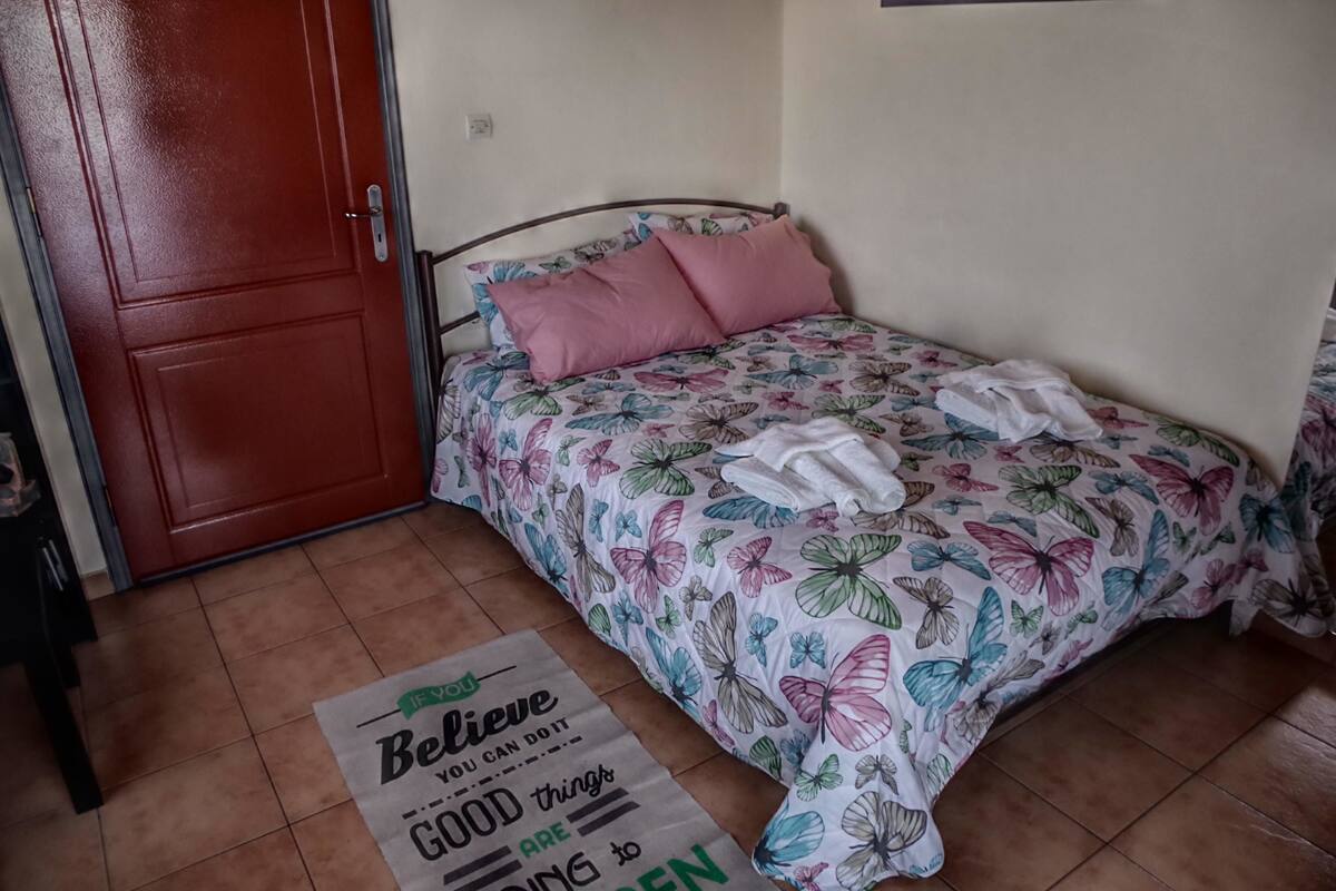 Ein Schlafzimmer mit einem Bett, das mit einer blumigen Tagesdecke bezogen ist, zwei rosafarbene Kissen und ein Paar weiße Handtücher. Ein motivierendes Zitat liegt auf dem Boden.