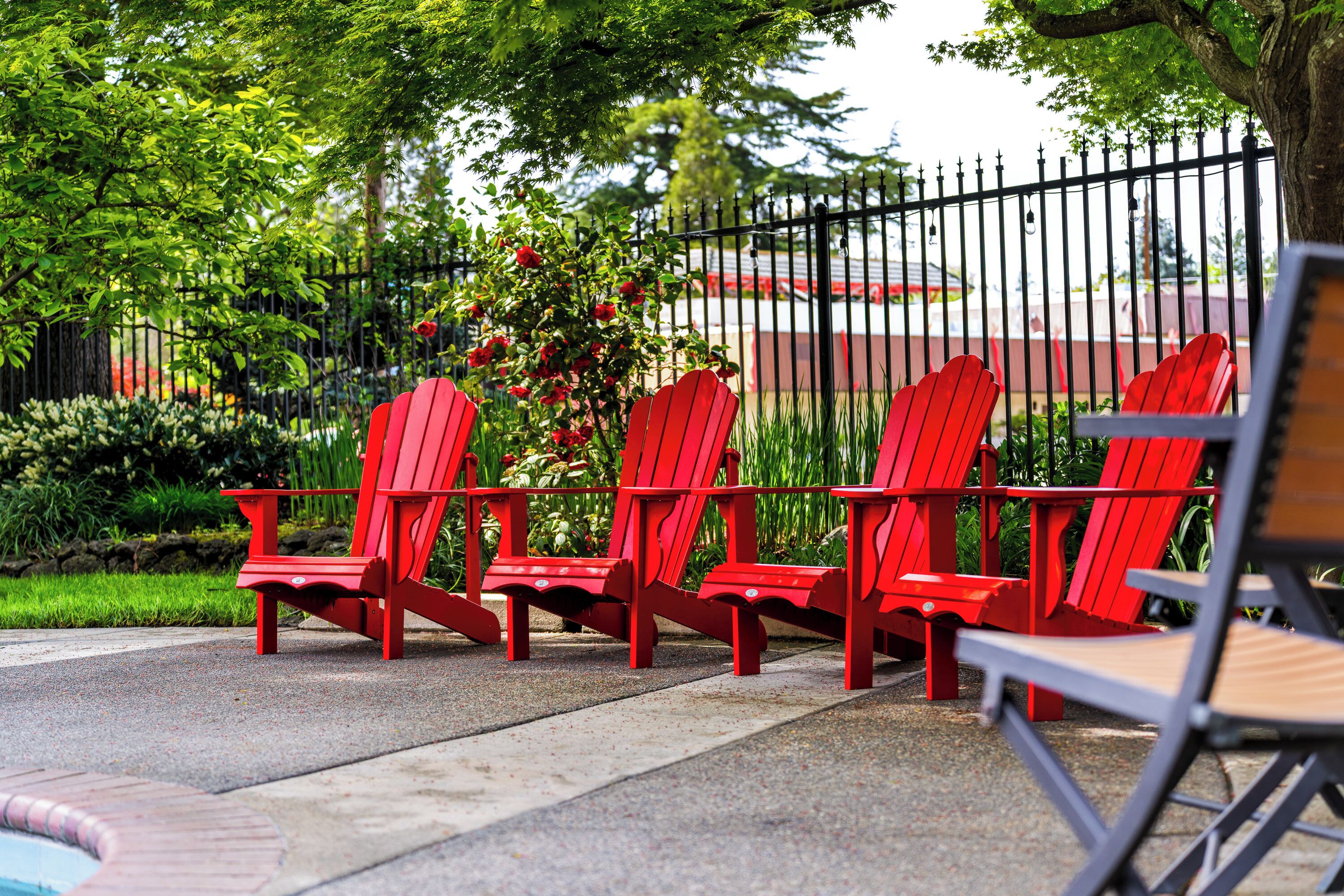 A row of red Adirondack chairs by a poolside, with a black fence and greenery in the background.