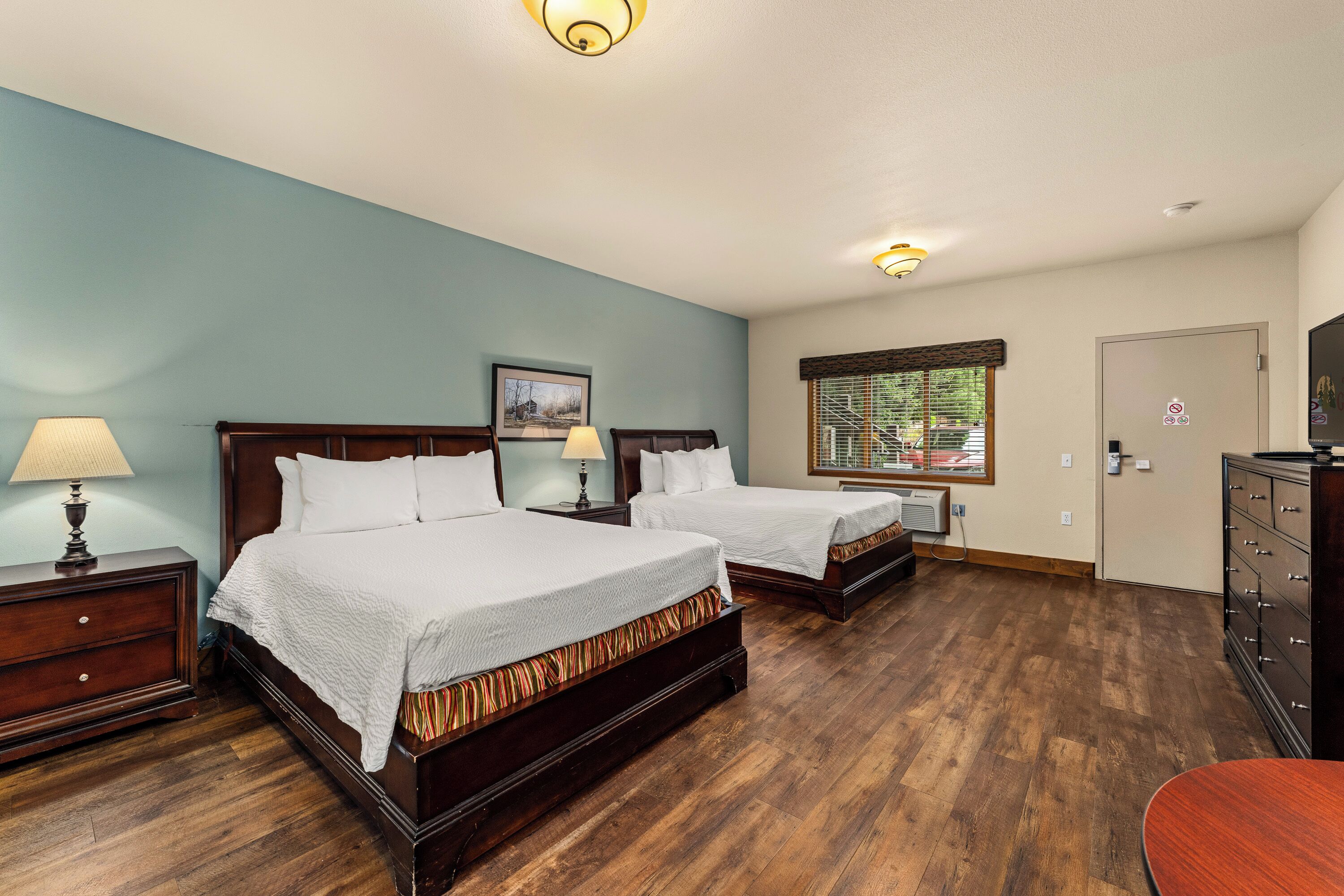 A hotel room with two beds, wooden floors, a dresser, a desk with a lamp, a window with blinds, and a door with a lock.