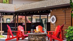 A patio area with a stone fire pit surrounded by red Adirondack chairs, a wooden fence, and a lifebuoy hanging on the wall. - Redwood Garden Inn (Grants Pass)