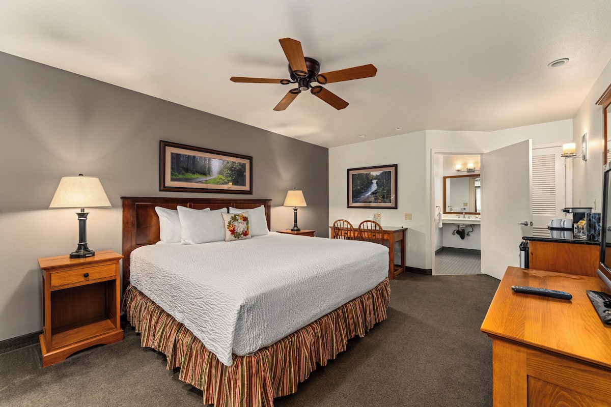 A hotel room with a bed, two bedside tables with lamps, a ceiling fan, a desk with a chair, a mirror, and framed pictures on the walls.
