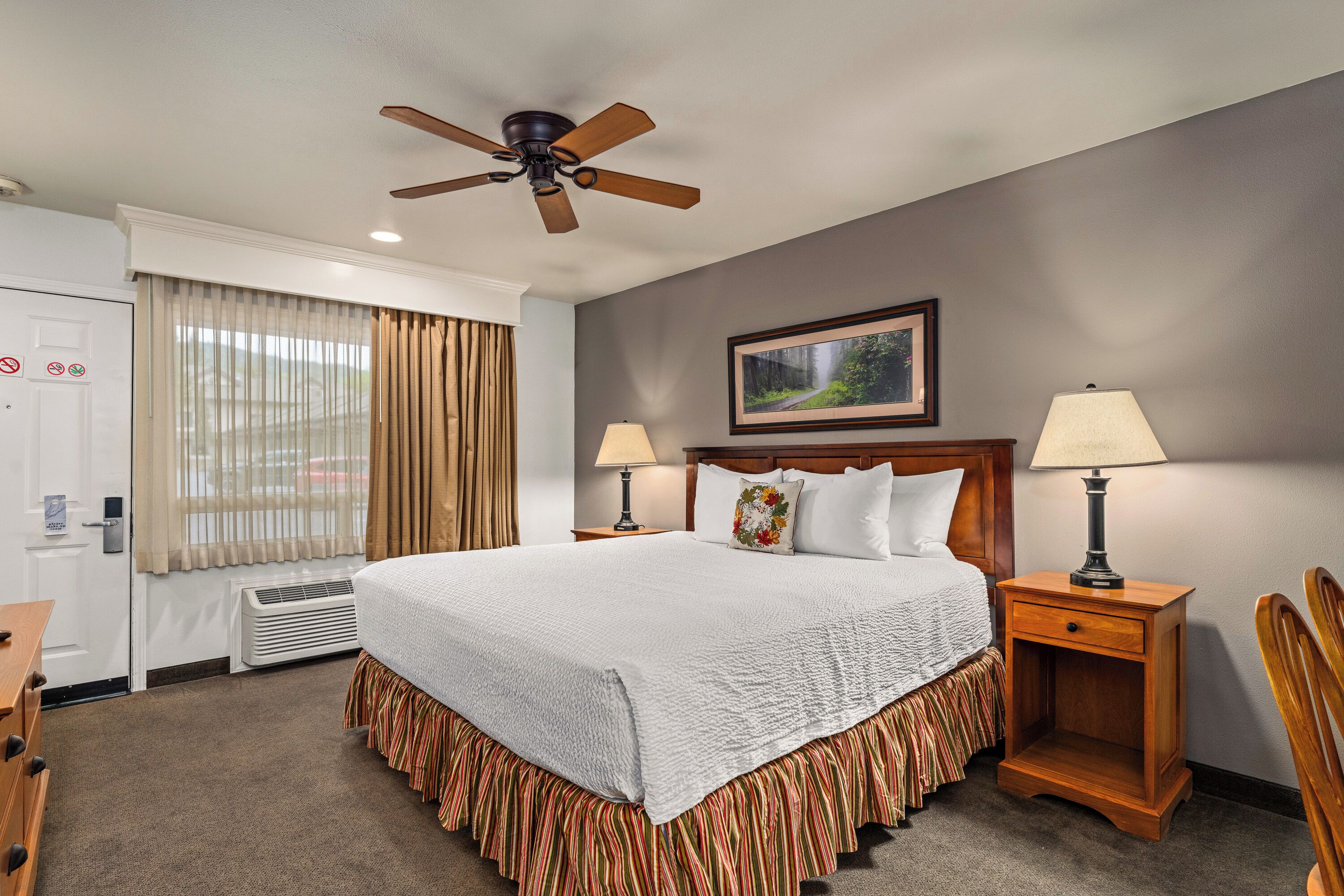 A hotel room with a bed, two bedside tables with lamps, a ceiling fan, a window with curtains, a framed picture on the wall, and a door with a sign.