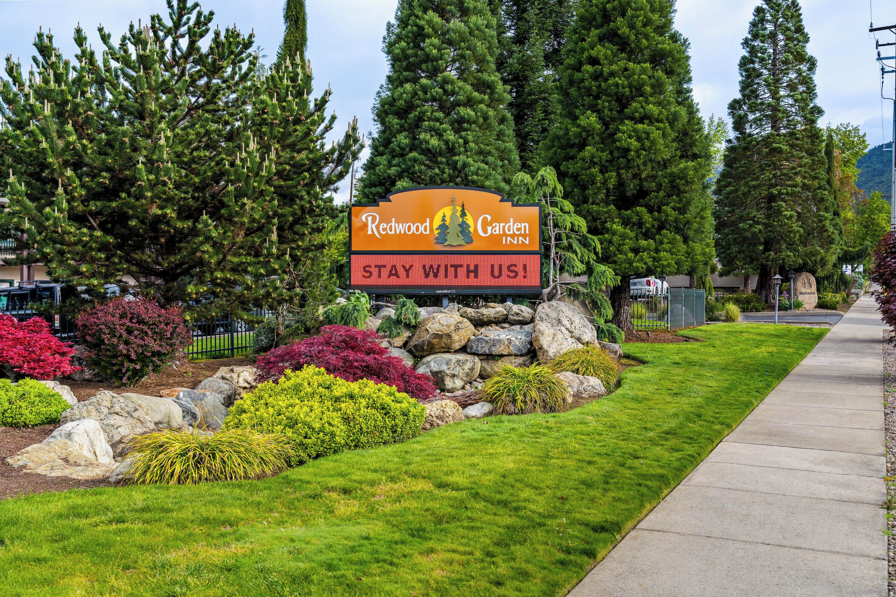 A sign for Redwood Garden Inn with a 'Stay with us!' message, surrounded by landscaped greenery and a paved walkway.