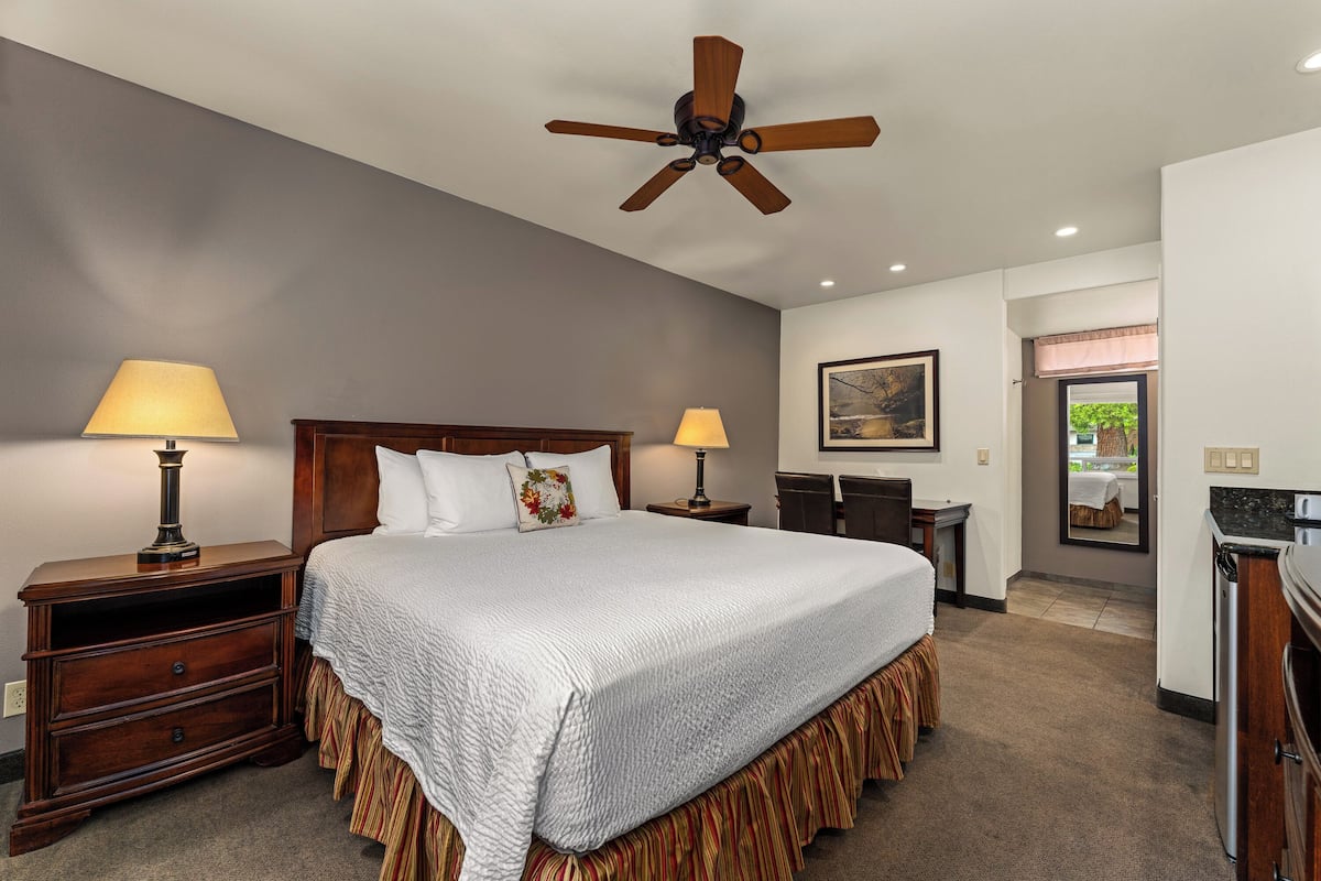 A bedroom with a bed, two bedside tables with lamps, a ceiling fan, a painting on the wall, and a view of the outside through a window.