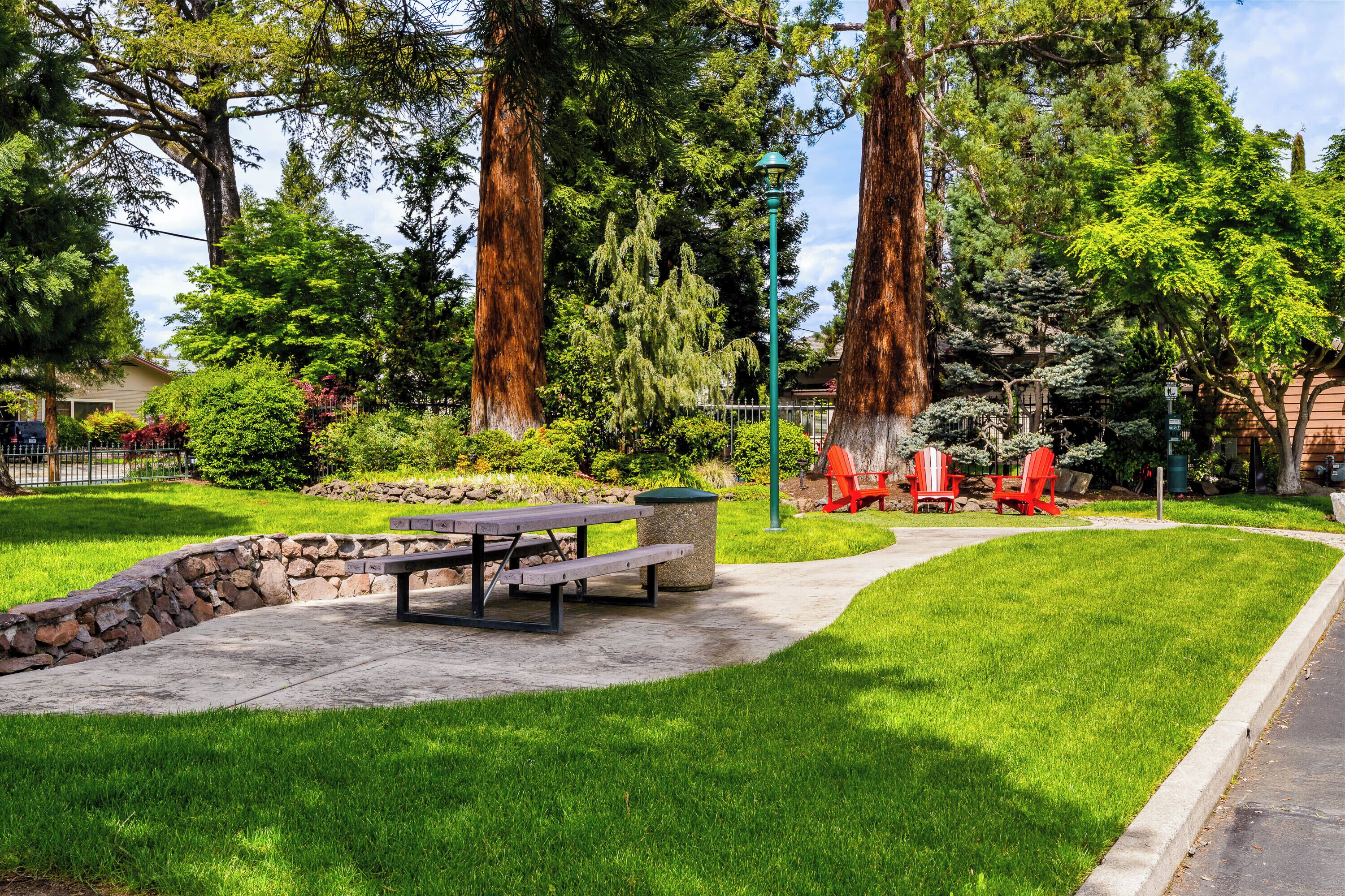 Un espace pique-nique équipé d’un banc, d’une table à pique-nique et de chaises rouges, le tout entouré d’une végétation luxuriante et de grands arbres.