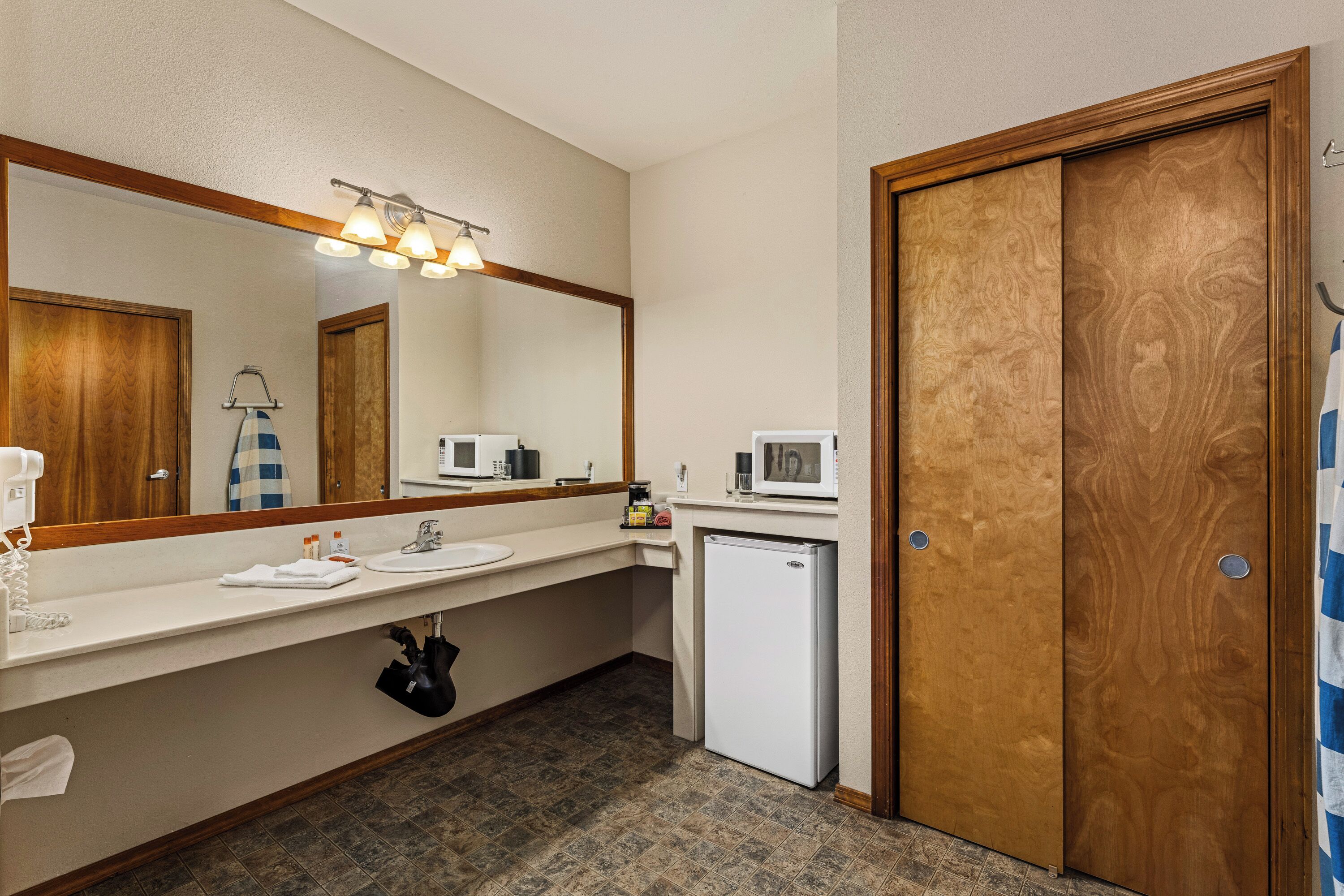 A hotel bathroom with a large mirror, a sink, a microwave, a refrigerator, and a wooden door.