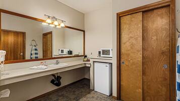 A hotel bathroom with a large mirror, a sink, a microwave, a refrigerator, and a wooden door.