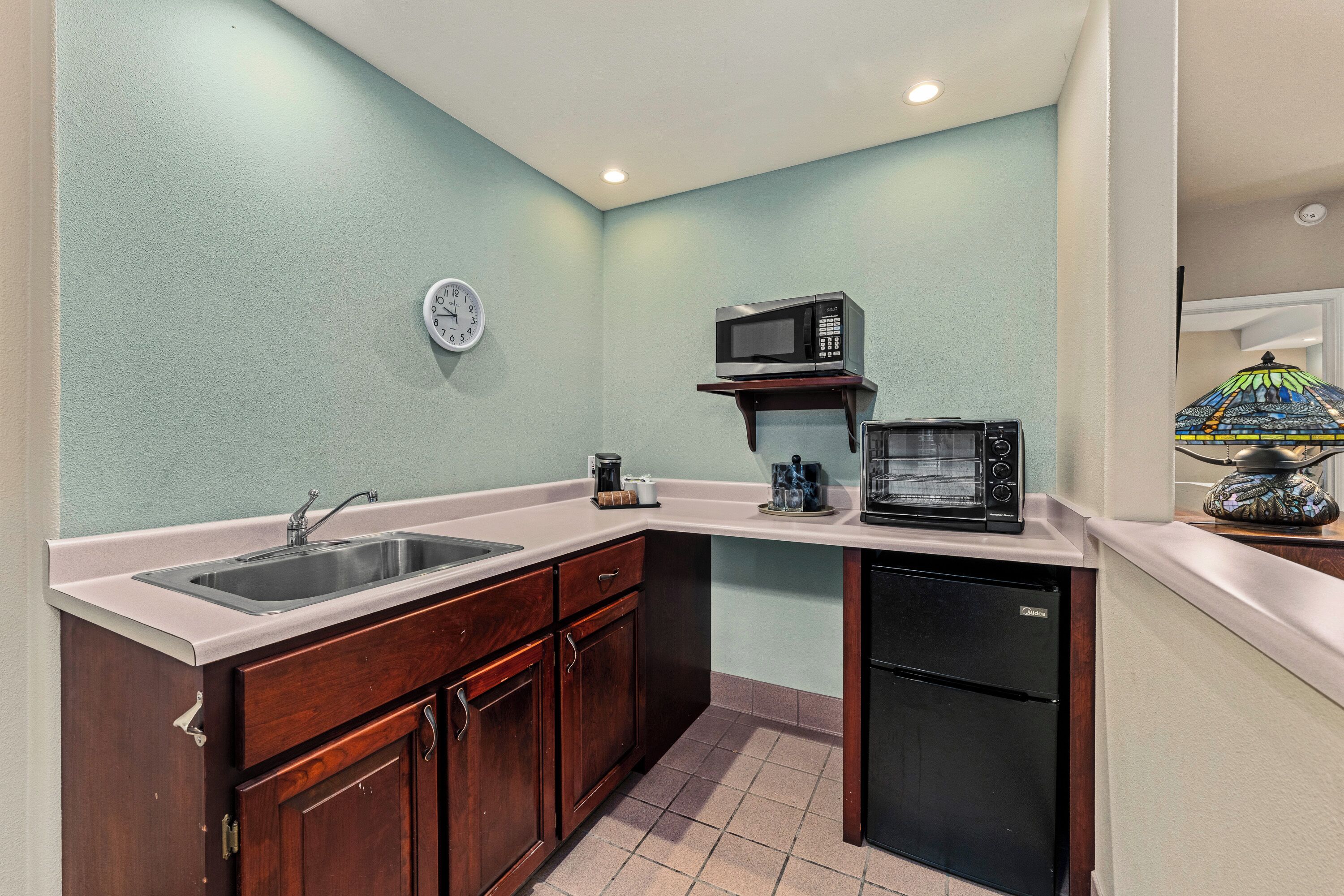 A compact kitchen with a sink, microwave, and refrigerator, wooden cabinets, and a clock on the wall.