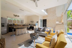 Una cocina moderna con una isla central, un comedor con taburetes de bar, una sala de estar con sofá y mesa de centro, y una escalera que conduce a un espacio exterior.