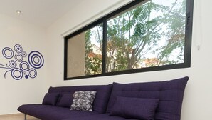 A modern room with a purple sofa, a decorative wall art piece, and a large window with a view of trees. - Prana 212 by BRM (Tulum)