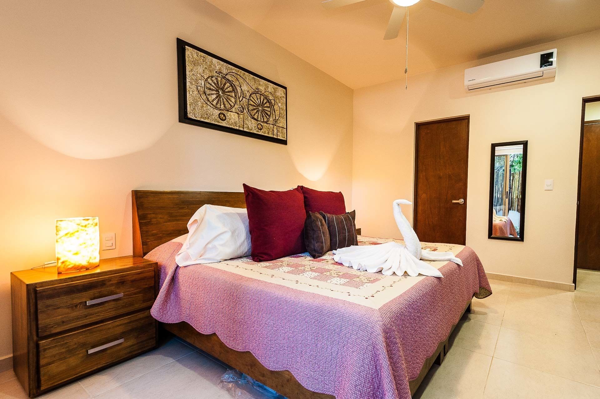 A neatly arranged bedroom with a wooden bed, a bedside table with a lamp, a framed artwork above the bed, and an air conditioning unit on the wall.