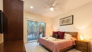 Une chambre à coucher comprenant un lit, une table de chevet, un ventilateur de plafond, une porte-fenêtre coulissante donnant sur un balcon et une œuvre d’art encadrée accrochée au mur.