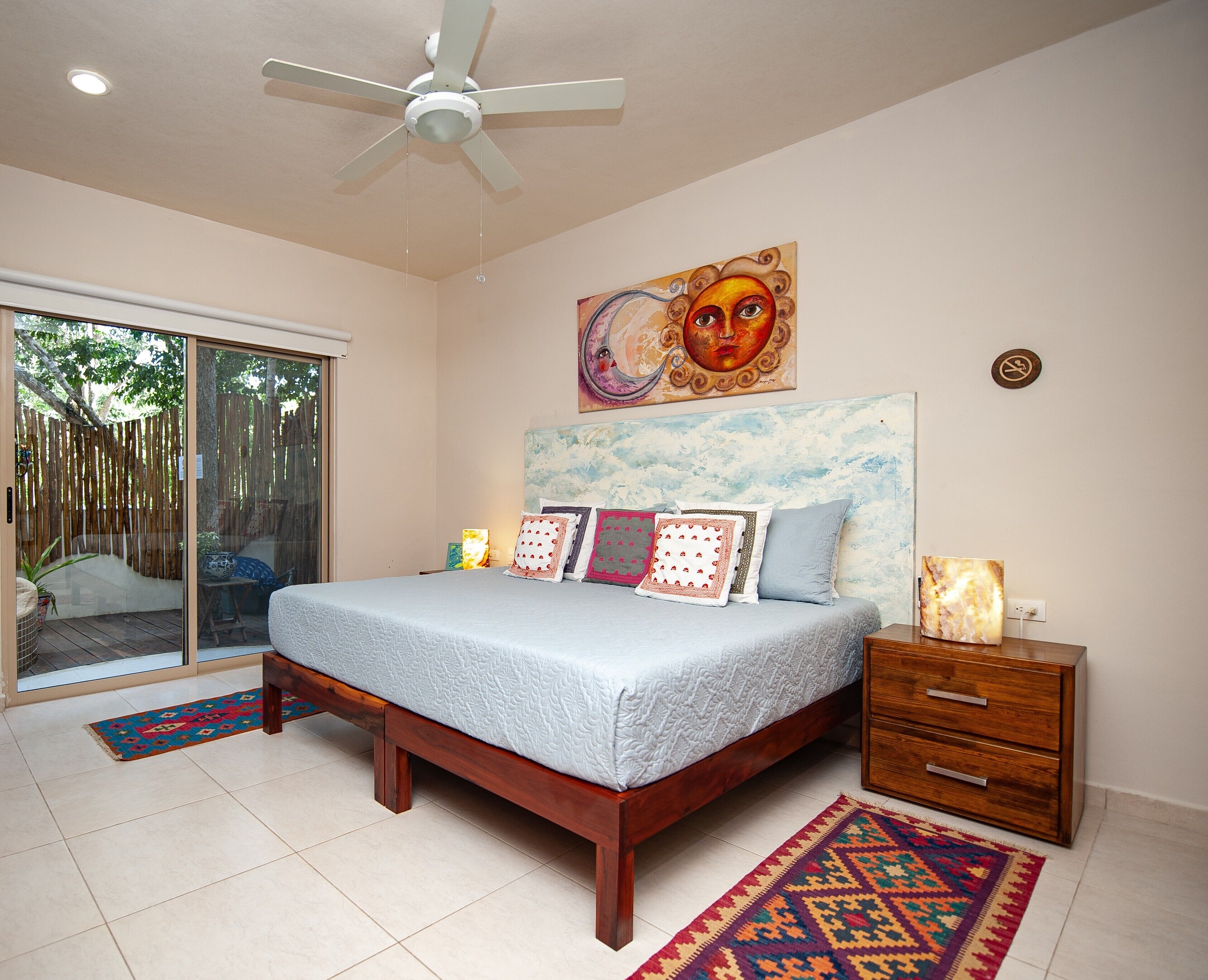 A bedroom with a bed, a nightstand, a ceiling fan, a painting, and a view through a sliding door.