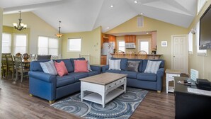 A spacious living area with a blue sectional sofa, a wooden coffee table, and a dining area with a table and chairs. - Dolphin Waves: Oceanside, Private Pool & Spa, Close to Beach, Quiet Neighborhood (Nags Head)