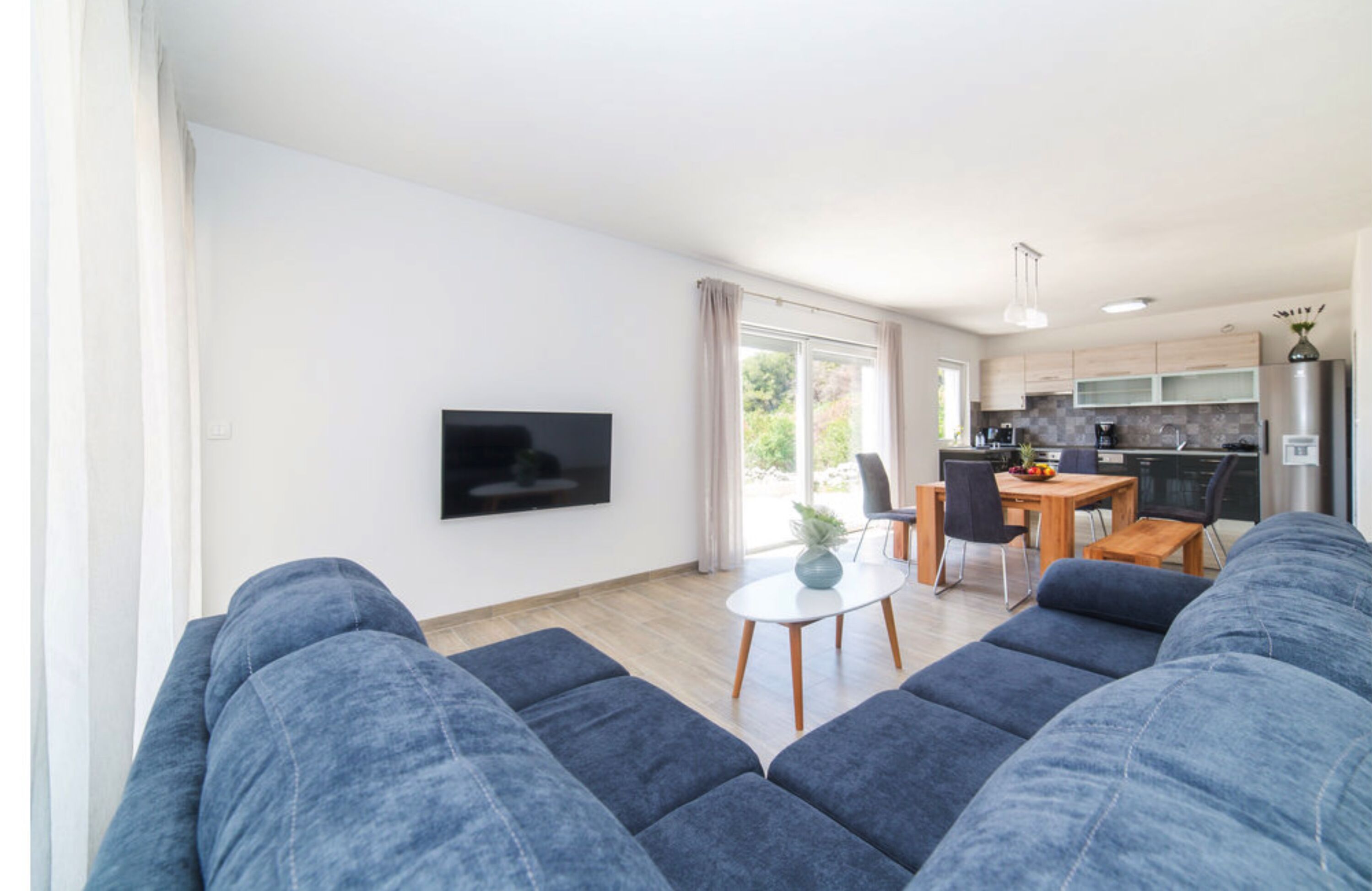 Una sala de estar moderna con un sofá grande, un televisor de pantalla plana montado en la pared, un comedor con mesa y sillas, y una cocina con refrigerador y armarios.