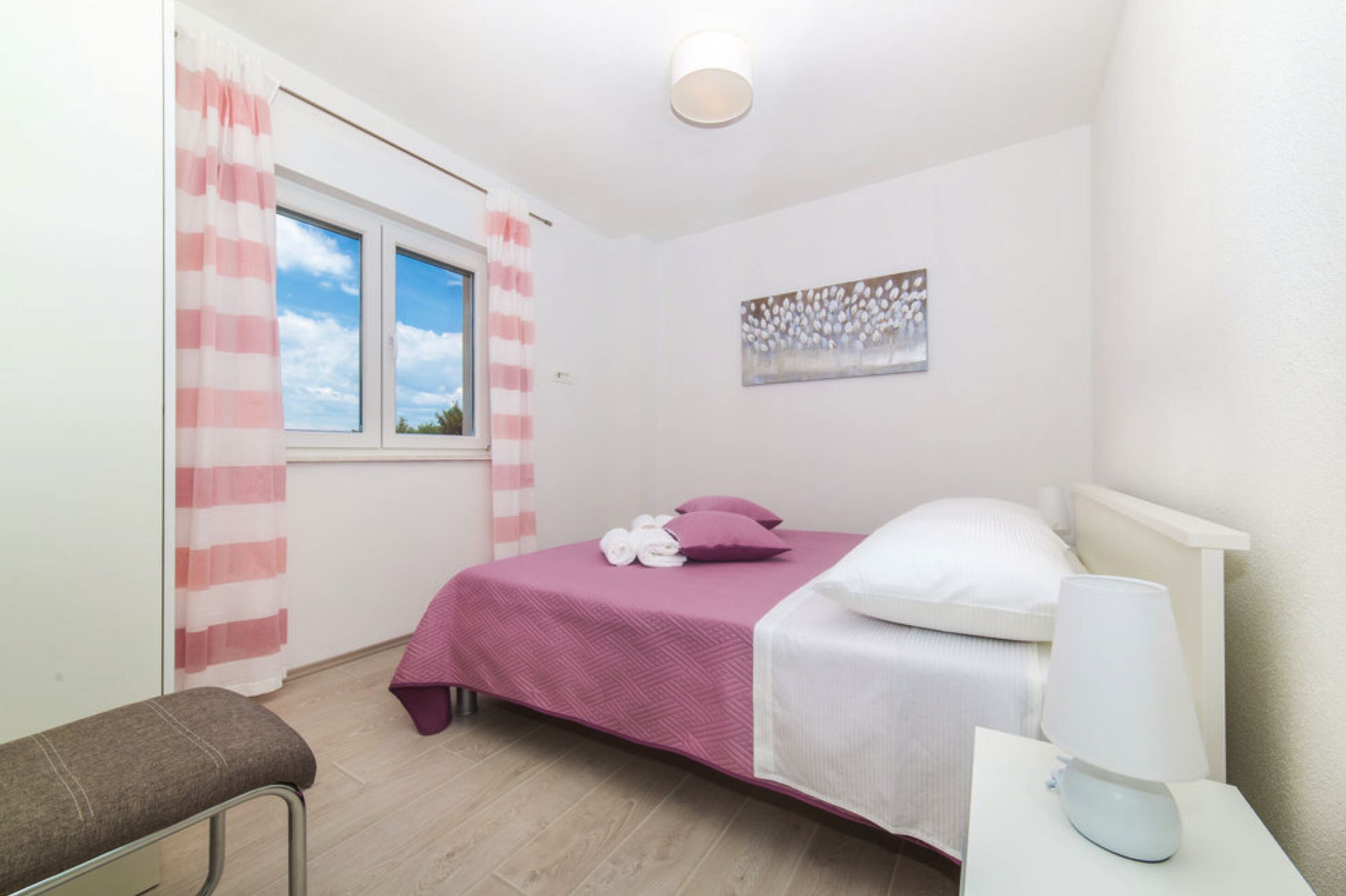 Un dormitorio con cama, un banco, una mesita de noche con lámpara, una ventana con cortinas y una obra de arte en la pared.