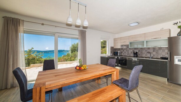 A modern kitchen with a large island, a dining area with a wooden table and chairs, and a view of the sea through large windows. - Holiday Home Goldy Trogir-Čiovo (Trogir)