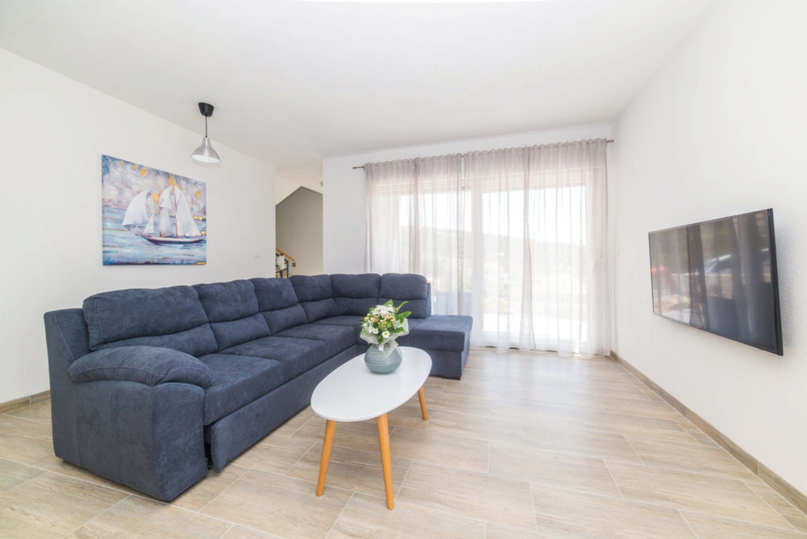 Una sala de estar moderna con un sofá seccional, una pequeña mesa blanca con un jarrón, un cuadro de un velero, un amplio ventanal con cortinas translúcidas y un televisor de pantalla plana montado en la pared.