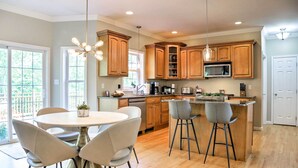 A modern kitchen with wooden cabinets, a central island with bar stools, a dining table with chairs, and a large window providing natural light. - Colonial-Style 'Warwick Retreat' - 1 Hour to NYC! (Monroe)