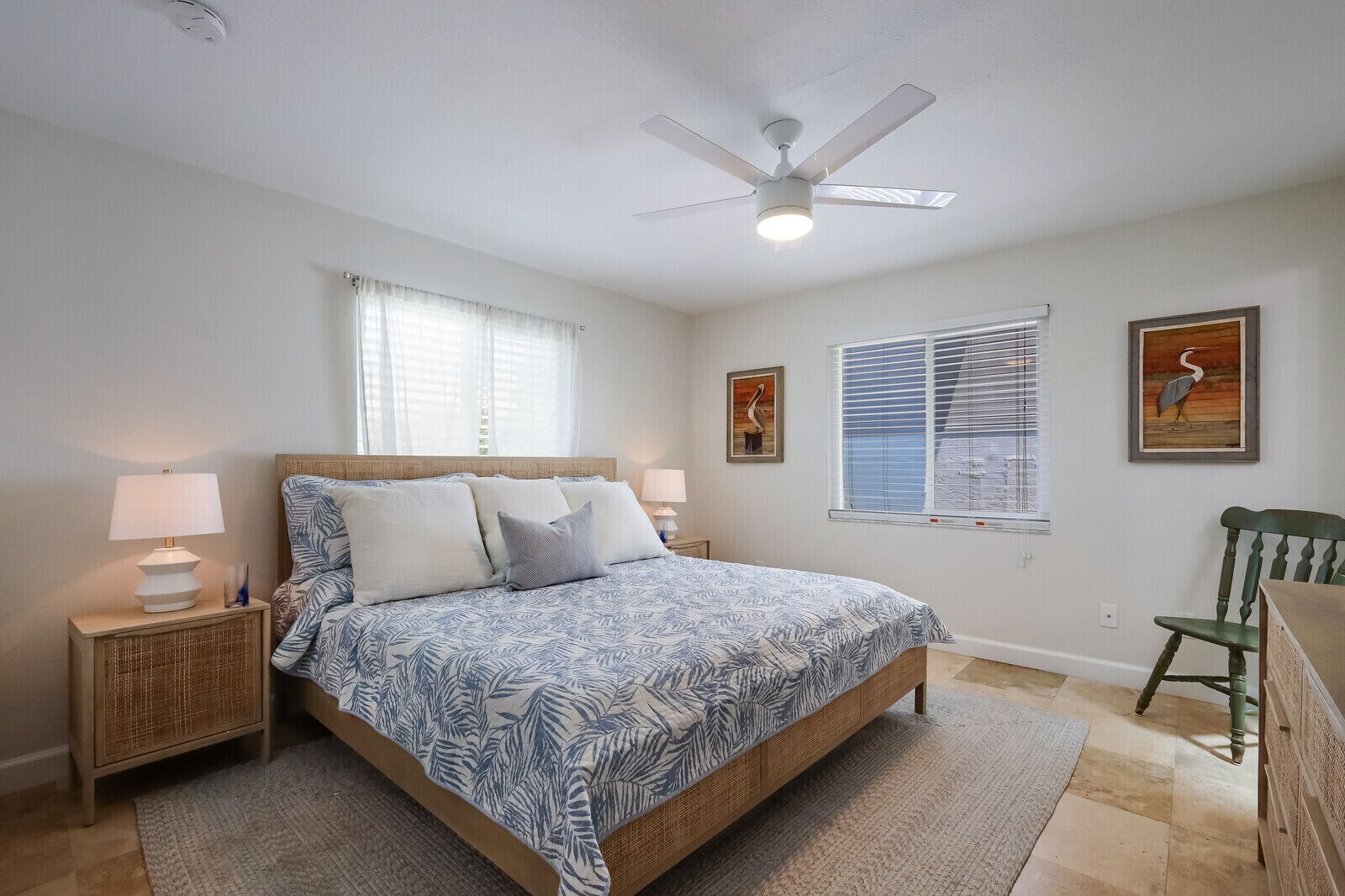 A bedroom with a bed, two bedside tables, a ceiling fan, a window with blinds, and two wall-mounted pictures.